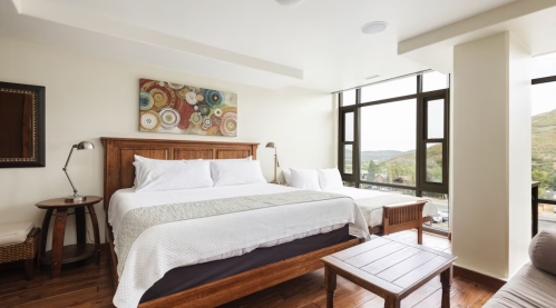 Bright, modern bedroom with a large bed, wooden furniture, and large windows showcasing a scenic view.